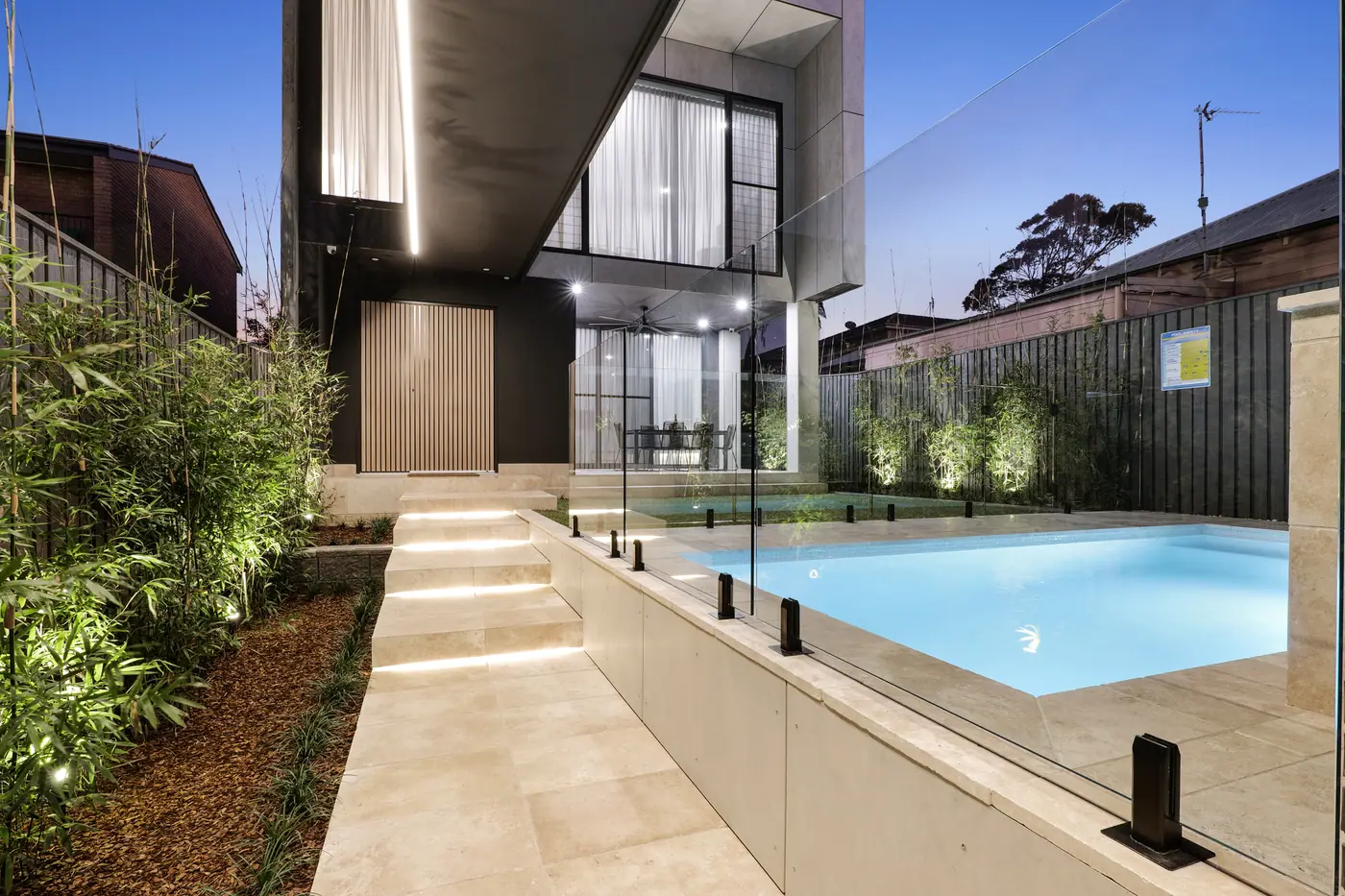 A plunge pool at dusk beside a two storey home, with lit stone steps running down to pale paving and frameless glass fencing around the water.
