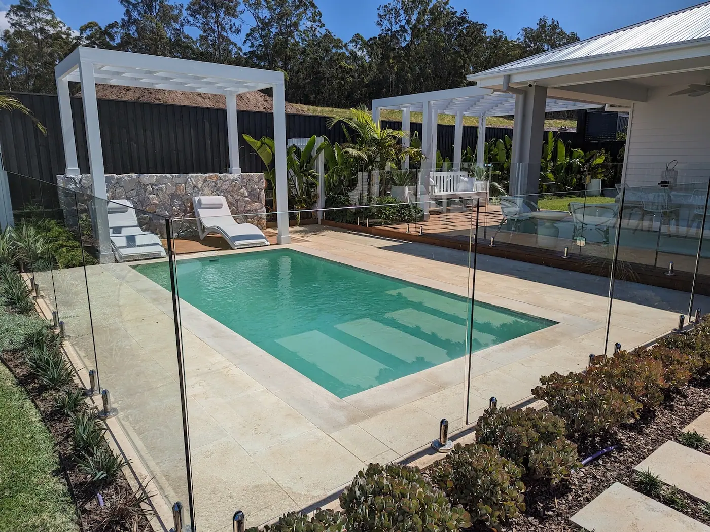 A green toned plunge pool between two white pergolas, with sun lounges on the paving and a stone clad garden wall behind.