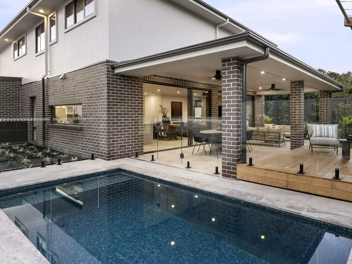 A dark tiled plunge pool reflecting a brick home with a covered alfresco area, glass fencing set into the stone coping at the water edge.