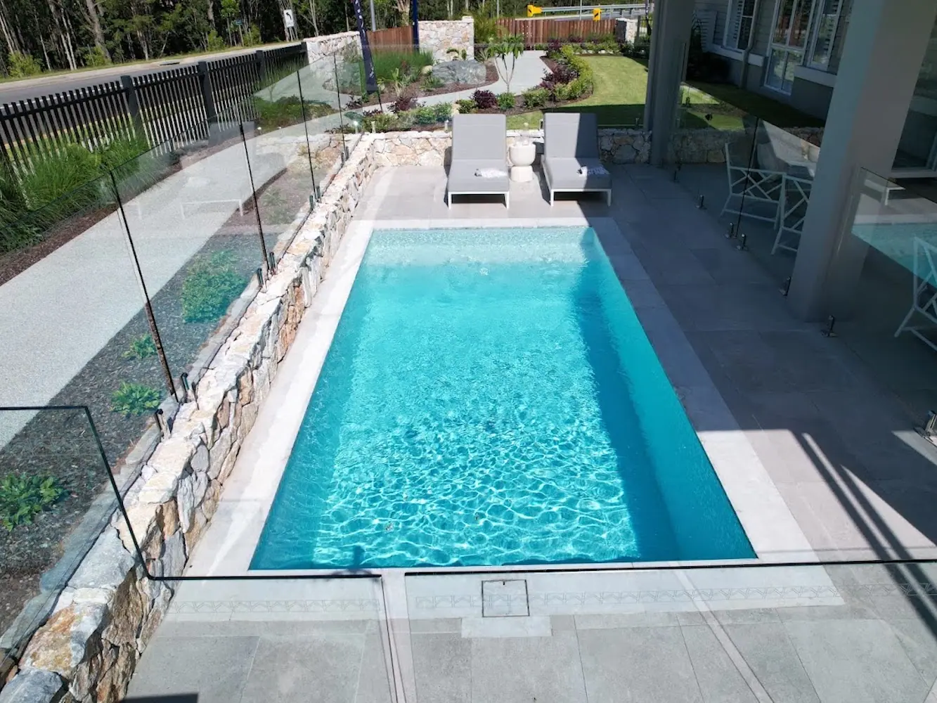 A plunge pool alongside a stone retaining wall, with sun lounges on the paving above it and glass fencing around three sides.