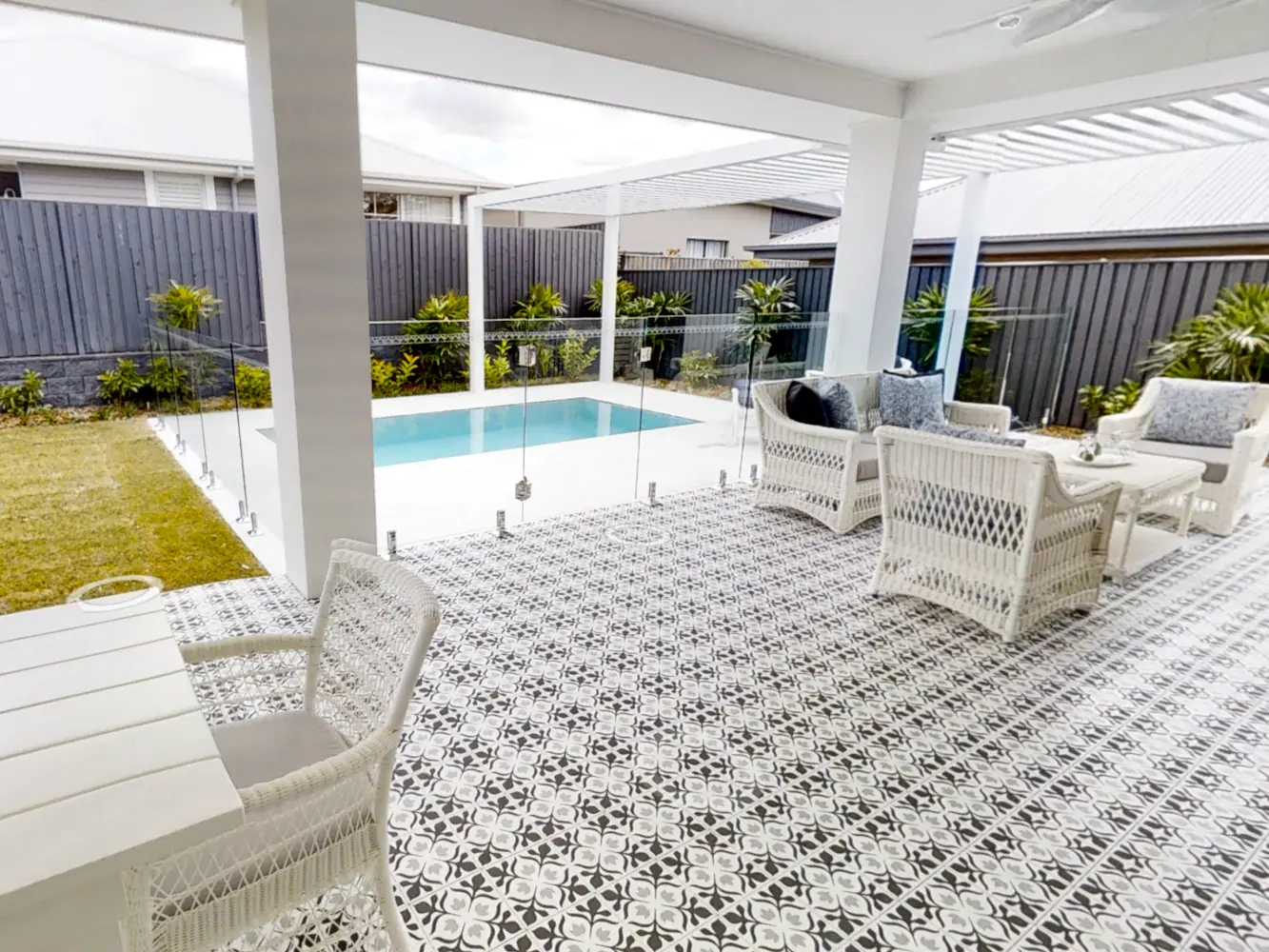A plunge pool seen from a covered patio laid with patterned floor tiles, white cane lounge chairs in the foreground and lawn beside the water.