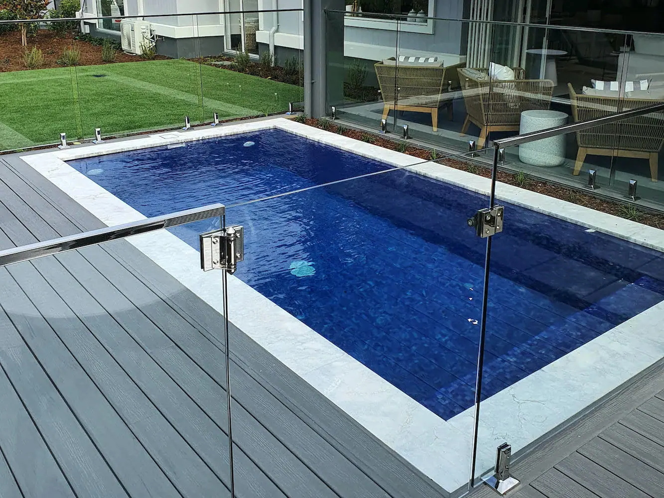 A deep blue tiled plunge pool set between grey composite decking and stone coping, with lawn and a glazed living area beyond the glass fence.
