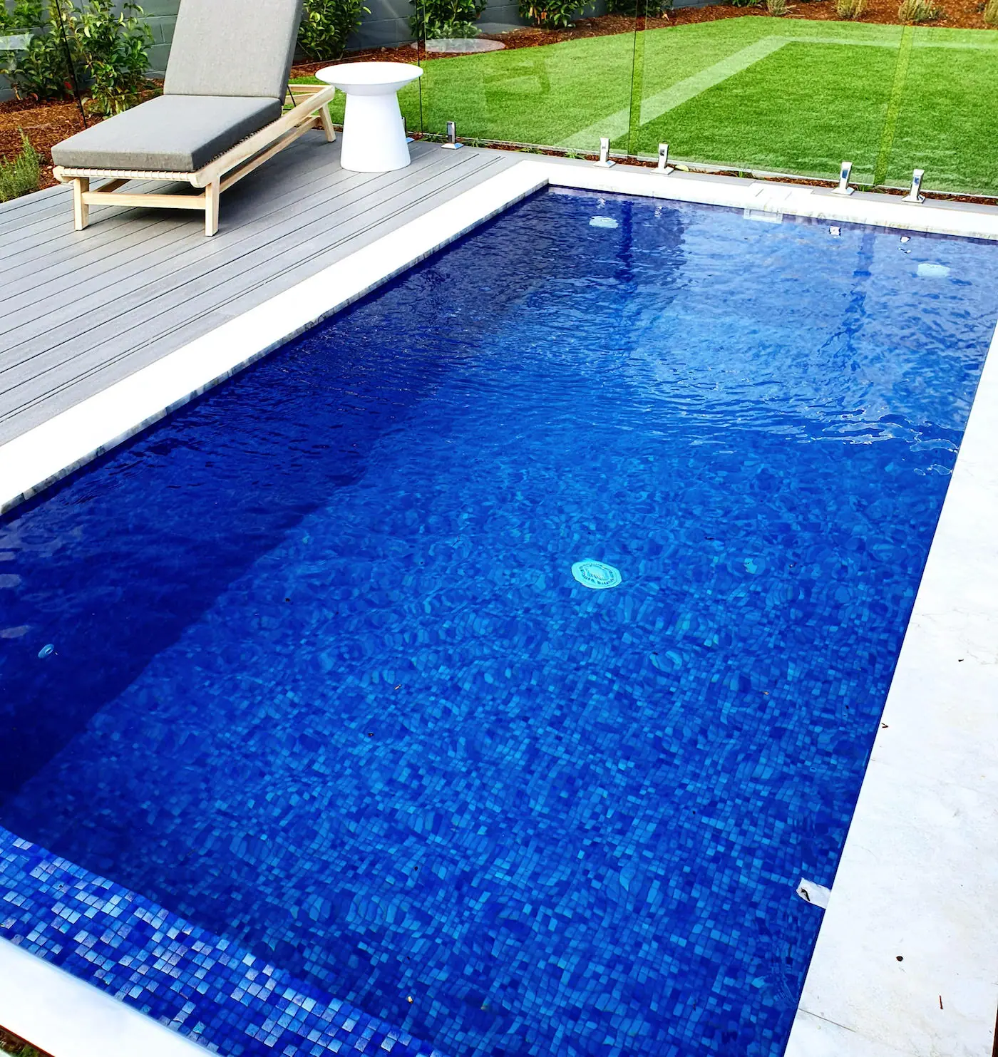 Looking down the length of a deep blue mosaic tiled plunge pool, with composite decking and a sun lounge on one side and lawn on the other.