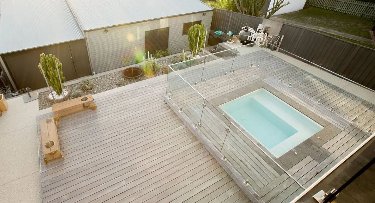 A compact plunge pool set flush into a large timber deck, with glass fencing around the water and cacti in a gravel garden bed alongside.