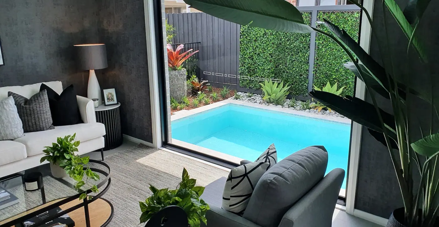 A plunge pool viewed from inside a living room through a wide opening, with a green plant wall and garden bed beyond the far edge of the water.
