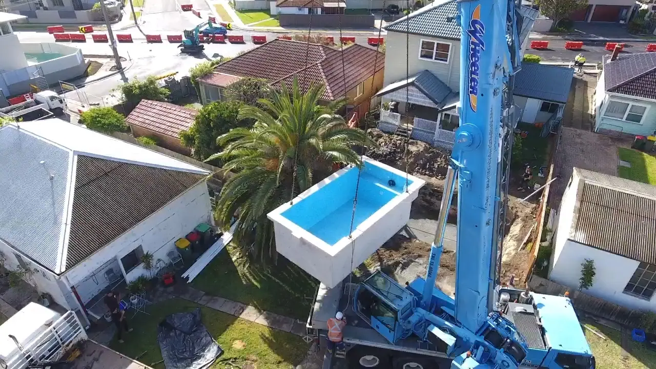 Aerial view of a mobile crane lifting a precast concrete pool shell high over the roofs of houses towards a backyard, with the street below closed off by traffic barriers.
