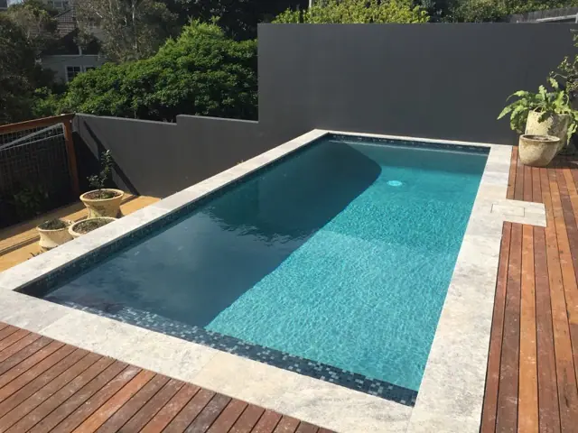 A plunge pool with a wide bench seat along one end and stone coping set into timber decking, against a rendered grey wall.