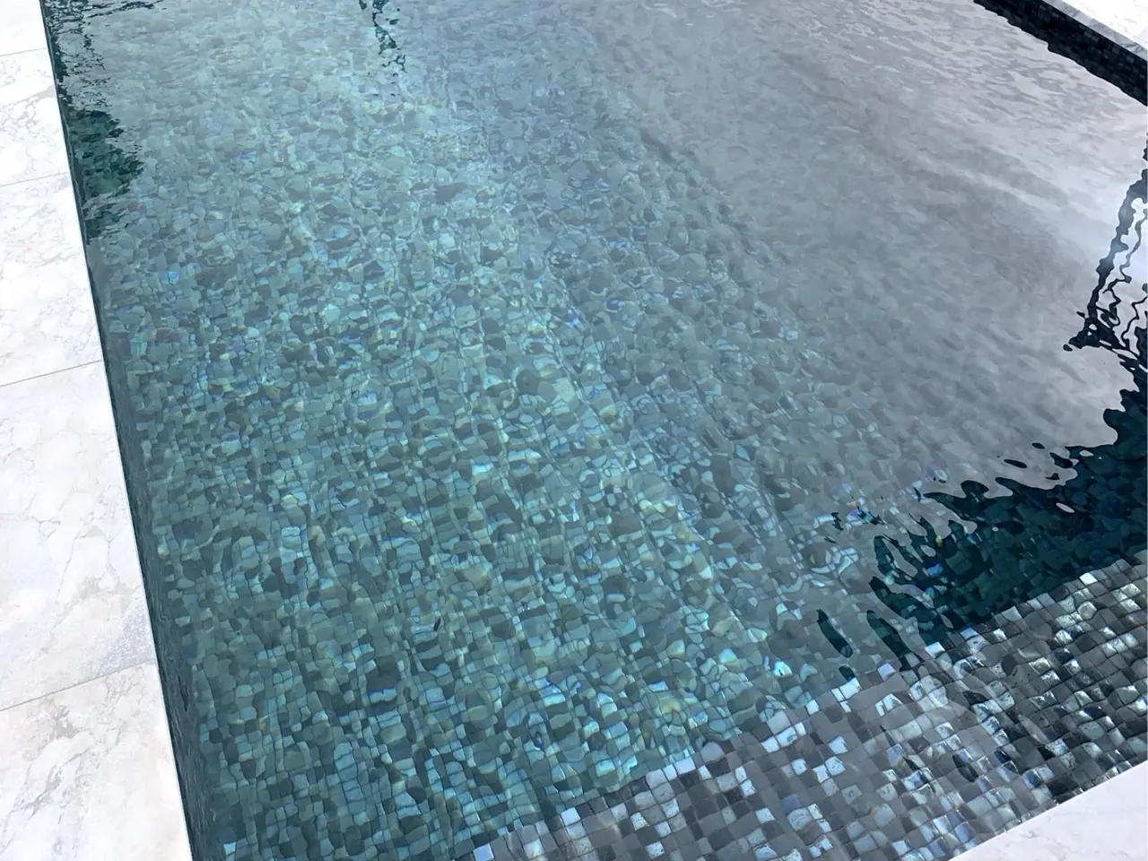 Close view of the finished water surface of a plunge pool over a blue green mosaic tiled interior, with pale stone coping at the edge.