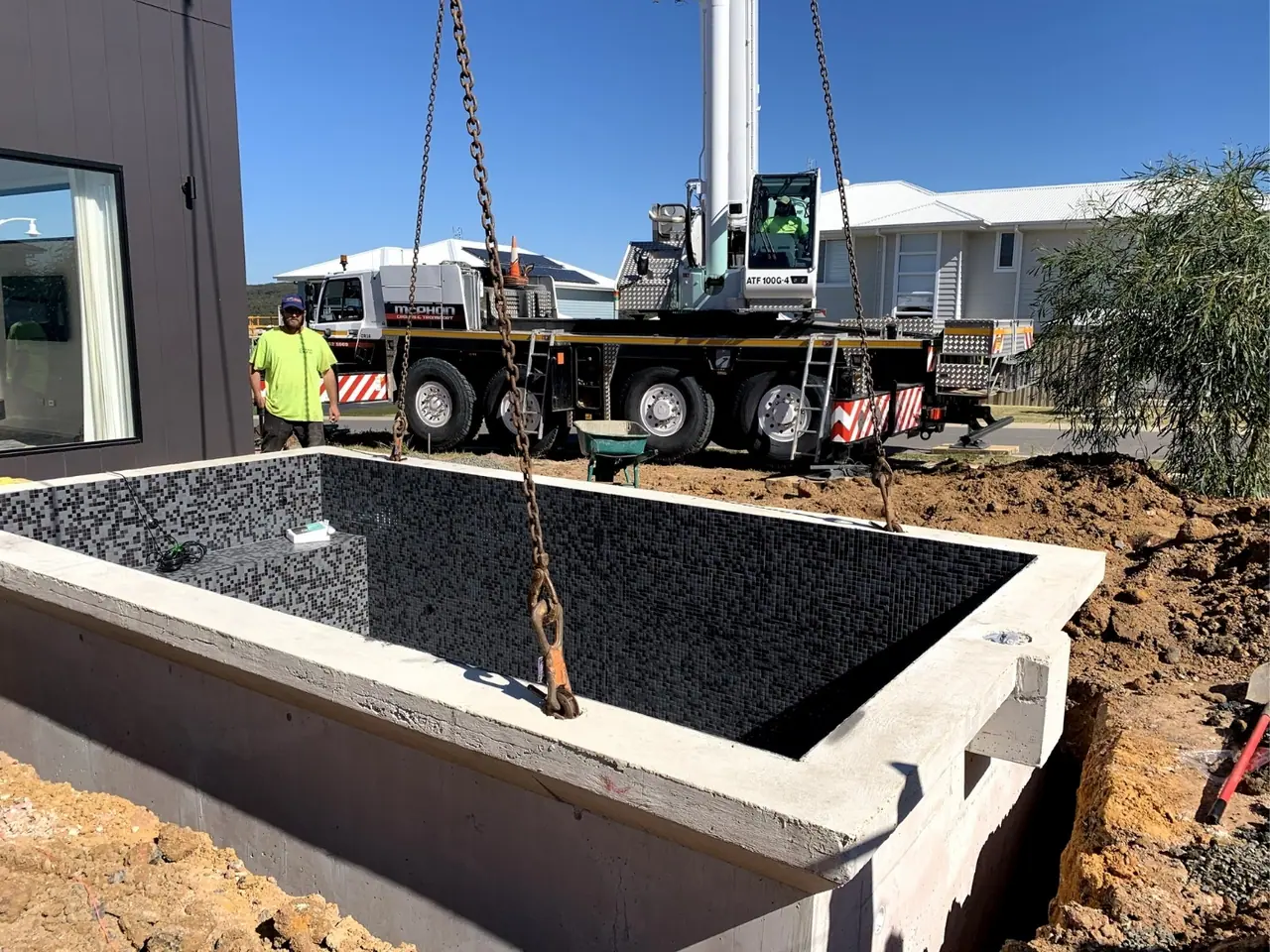 A precast concrete pool shell with a dark tiled interior hanging on chains from a mobile crane, being lowered into a prepared excavation beside a house.