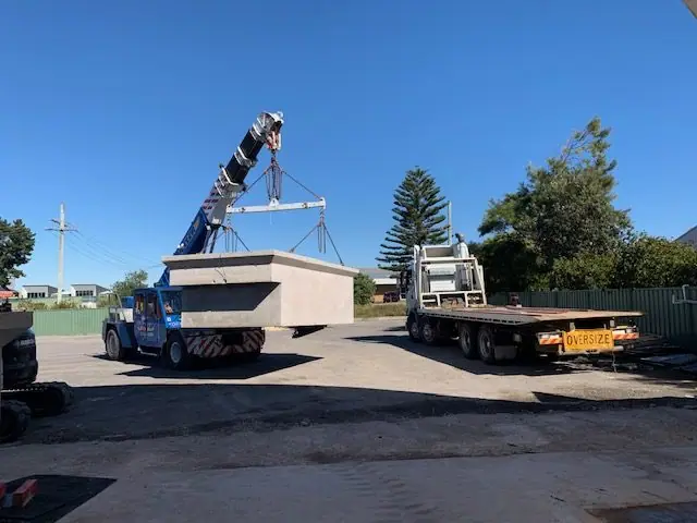 A mobile crane lifting a finished precast concrete pool shell across a yard towards a flatbed trailer marked as an oversize load.
